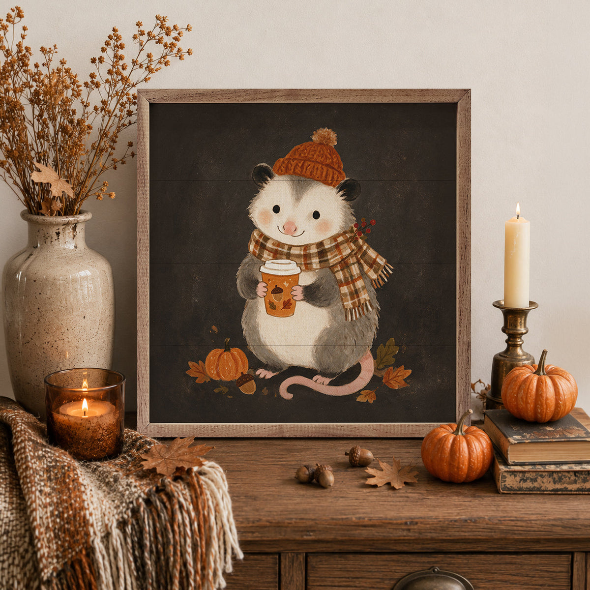 A framed autumn-themed art print featuring a cute gray and white raccoon wearing a brown knit hat and plaid scarf while holding a coffee cup, displayed on a wooden console table with pumpkins, a candle, dried flowers, and a throw blanket.