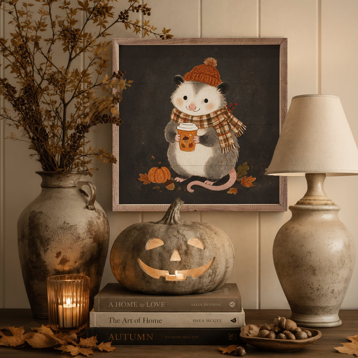 A framed autumn-themed art print featuring a cute gray possum wearing a brown knit hat and plaid scarf while holding an orange mug, displayed on a shelf with a carved pumpkin, books, a lamp, and dried branches.