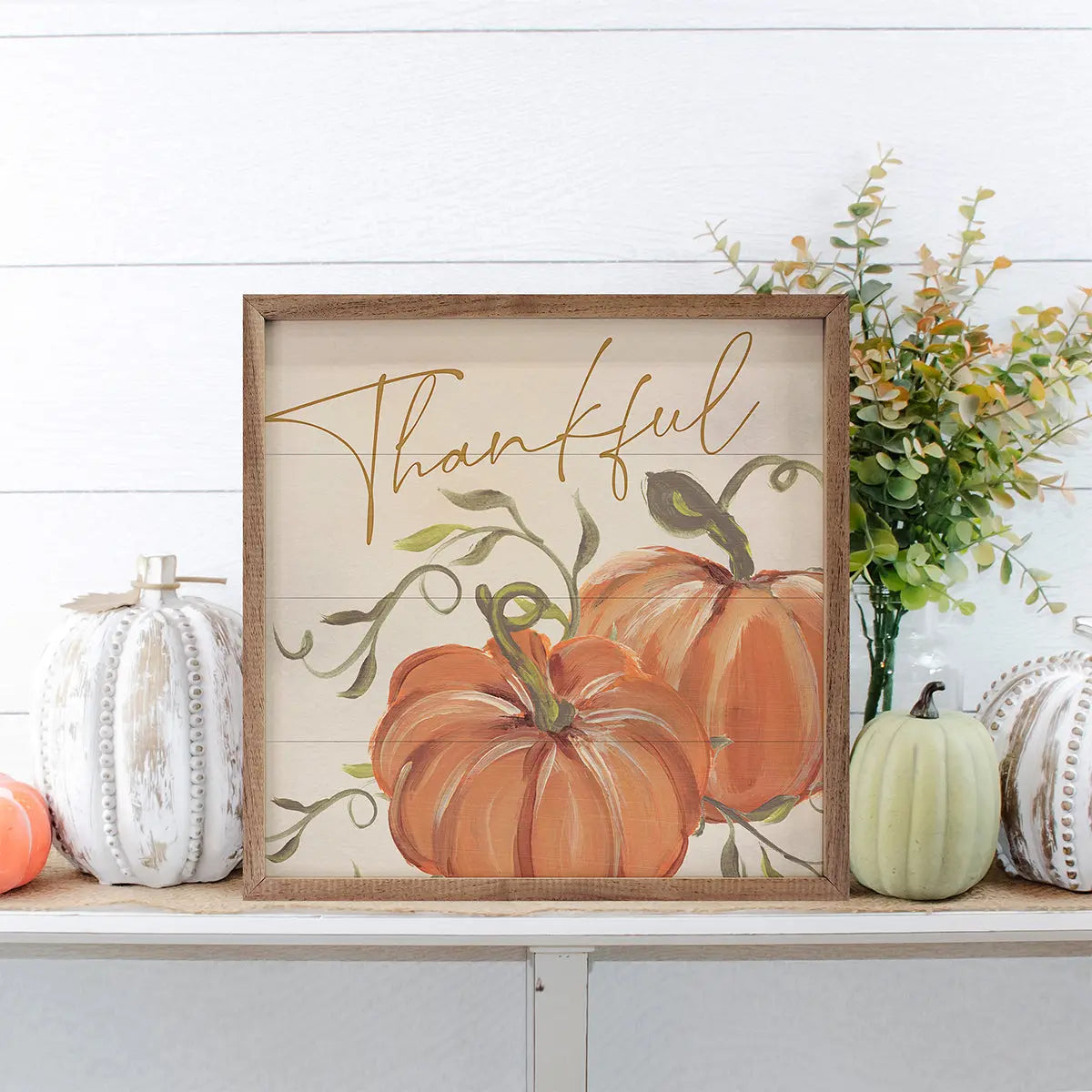 Decorative sign with pumpkins and 'Thankful' text on a shelf with pumpkins and flowers.