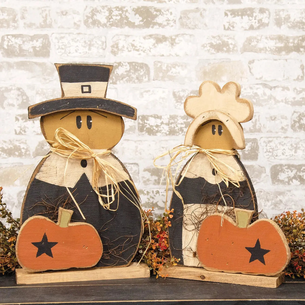 Two rustic wooden Thanksgiving decorative figures with black and beige striped bodies, straw neck bows, black hats, and orange pumpkin accents with black stars, displayed against a white brick wall.
