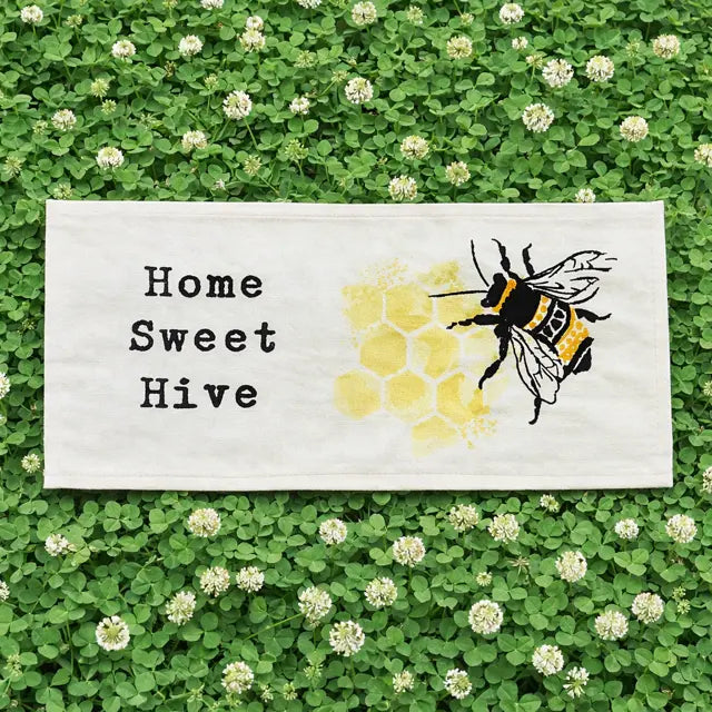 A rectangular white fabric sign or towel laid flat on a bed of green clover with small white flowers. The fabric has black text reading 'Home Sweet Hive' and a yellow honeycomb pattern with a black-and-yellow bumblebee illustration.