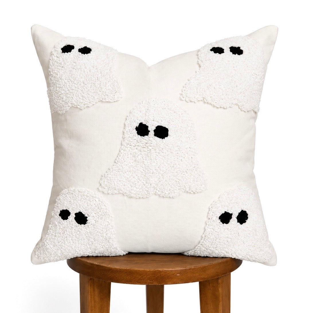 An off-white throw pillow with a textured, shaggy surface featuring four white ghost appliqués with black embroidered eyes, displayed on a wooden stool against a white background.