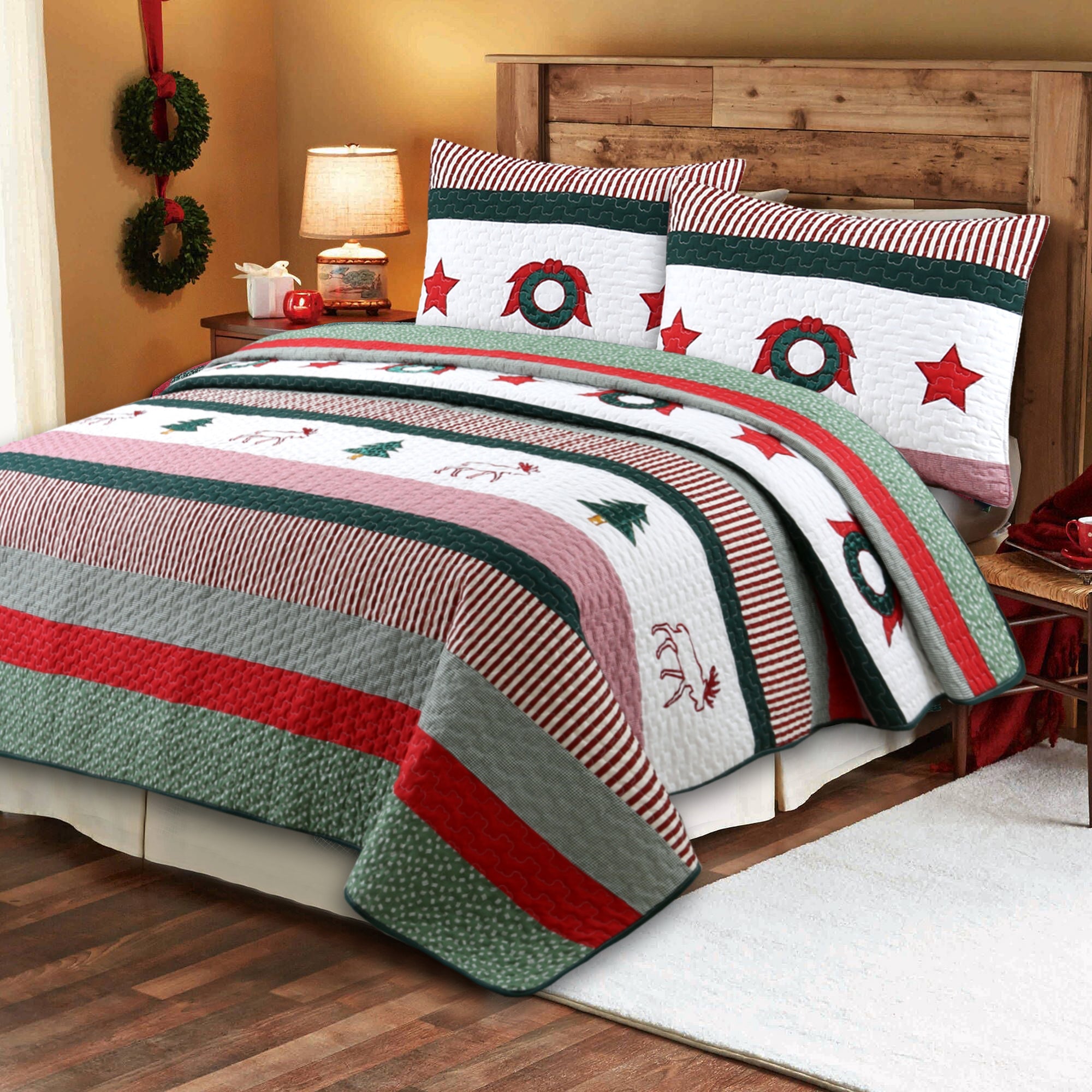 A Christmas-themed quilted bedding set with matching pillow shams, featuring stripes and motifs of green wreaths, red stars, pine trees, and snowflakes on a white background, styled on a wooden bed in a warm bedroom setting.