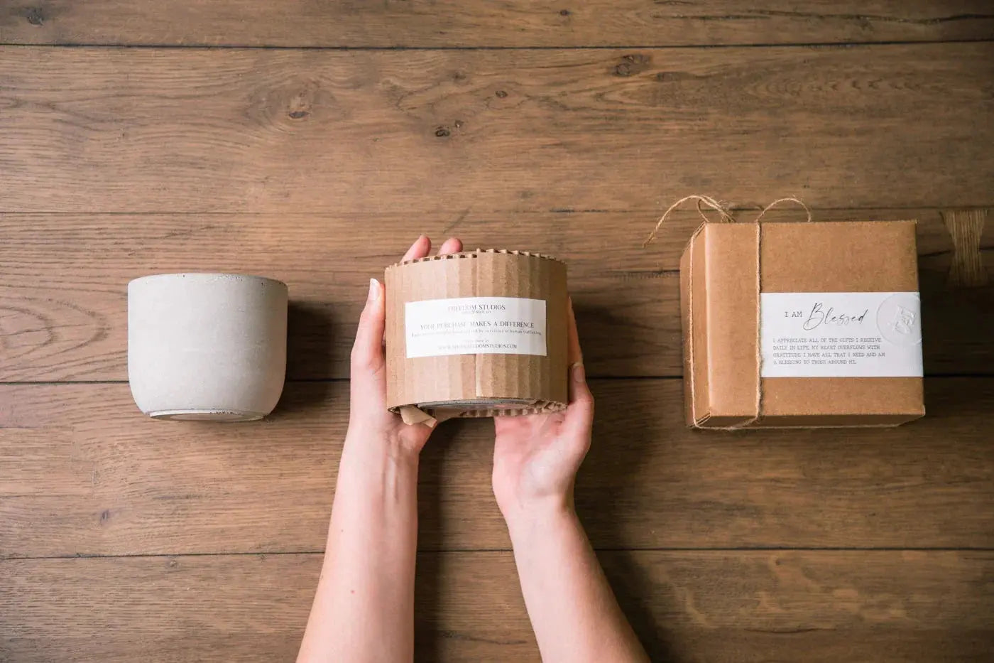 Hands holding a wrapped candle beside a ceramic cup and gift box on rustic wood, cozy home decor