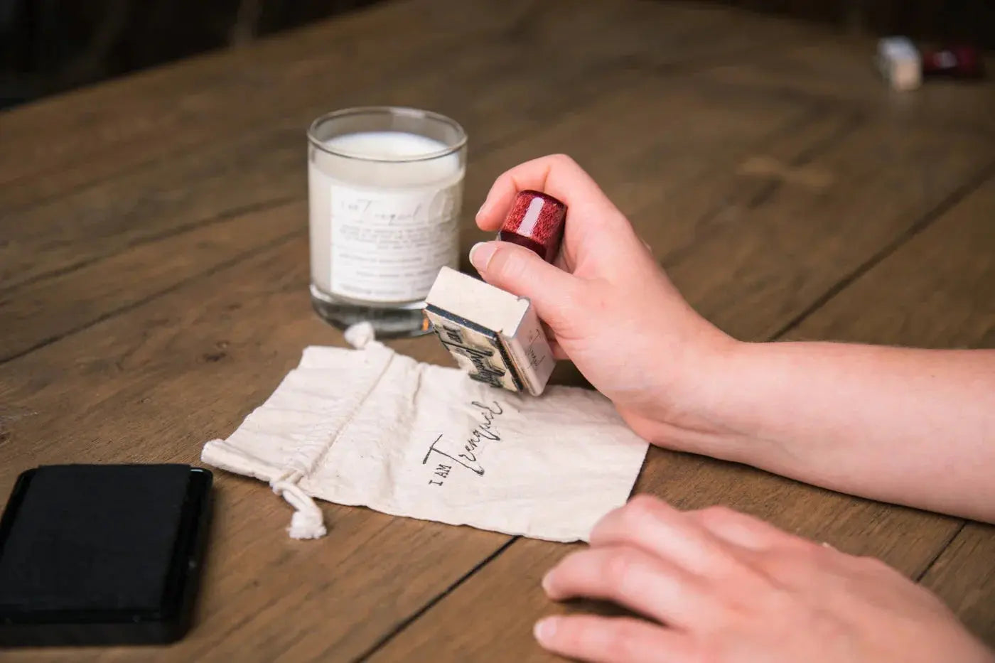 Hand stamping 'I Am Tranquil' on a cotton drawstring bag beside a soy candle, cozy home decor