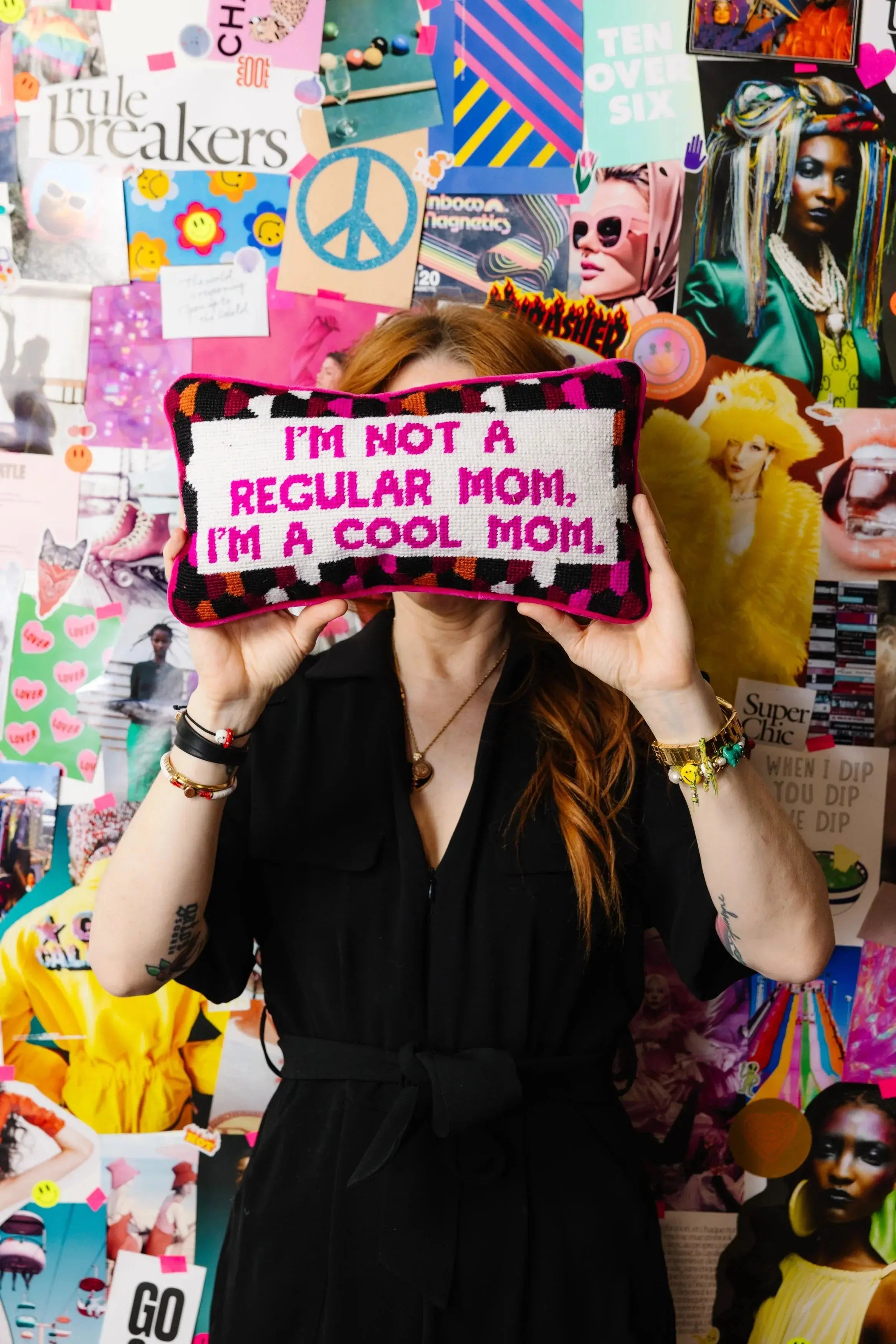 Cool mom needlepoint pillow with bold pink text, held against vibrant collage wall, perfect for quirky home decor.
