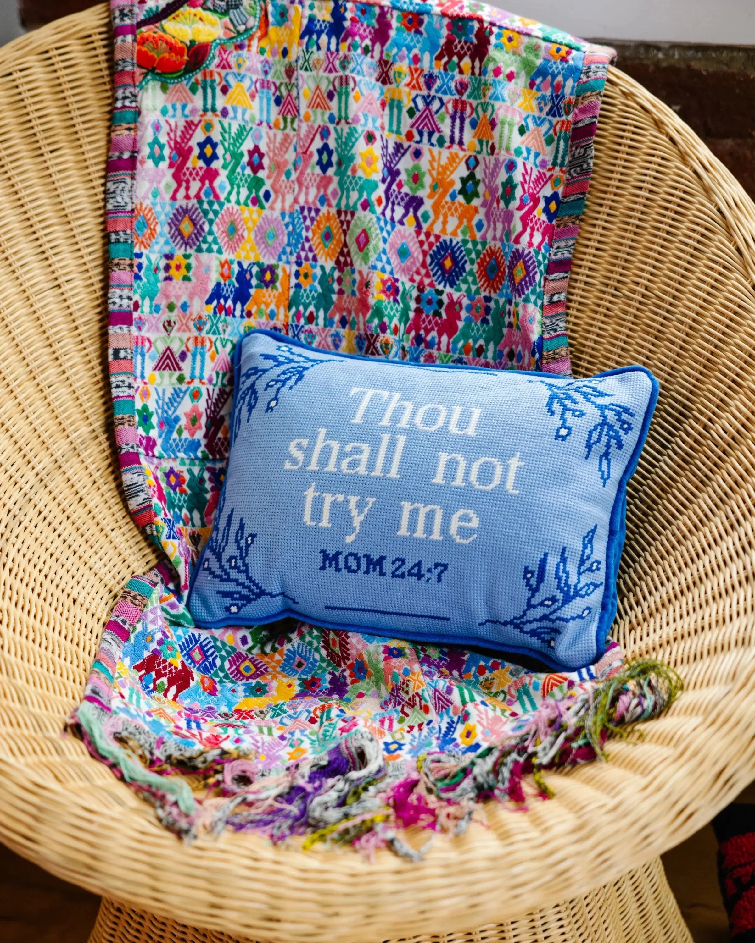 Blue embroidered pillow with 'Thou shall not try me MOM 24:7' on wicker chair with colorful patterned blanket