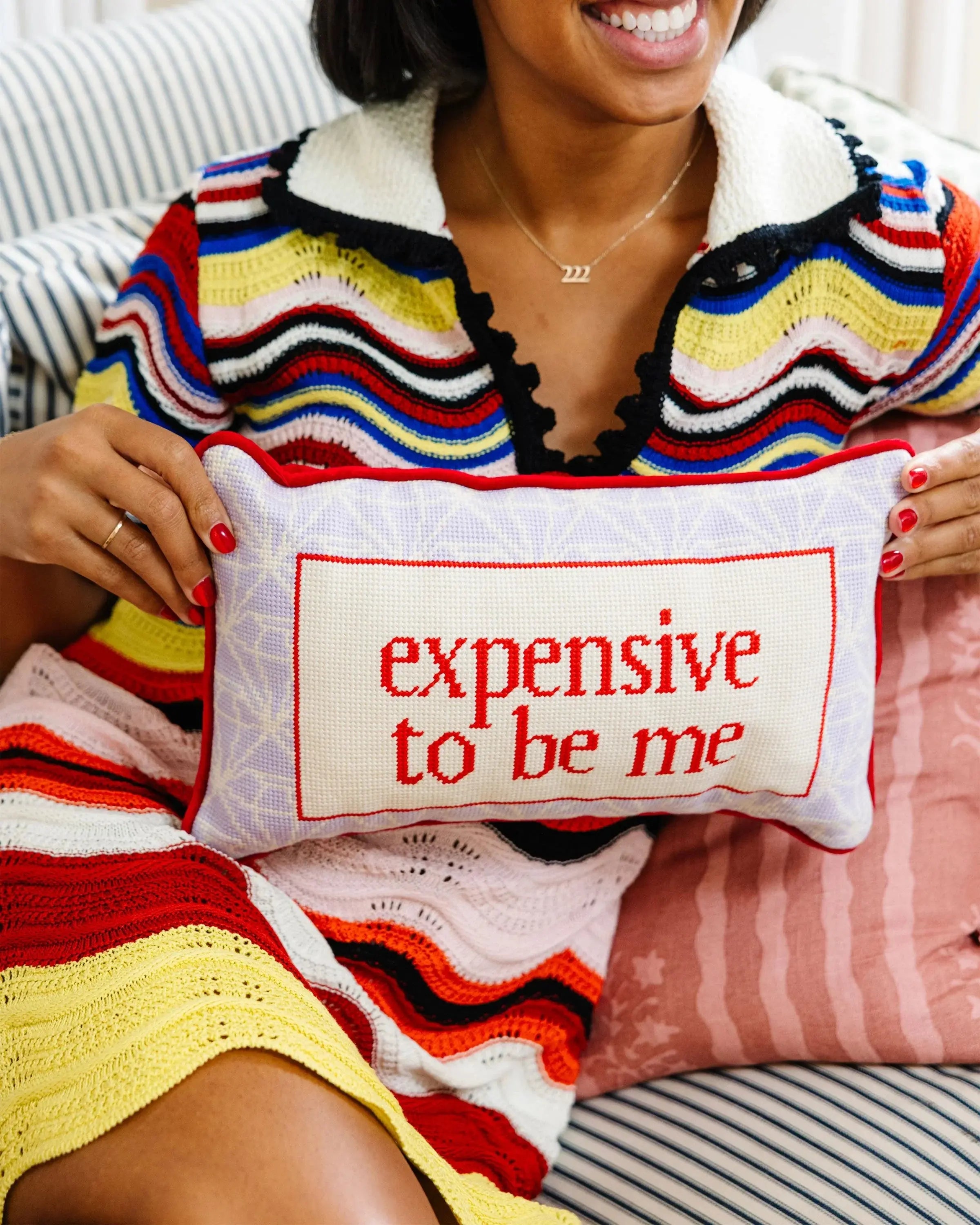 Person holding a pillow with 'expensive to be me' text, wearing a colorful sweater.