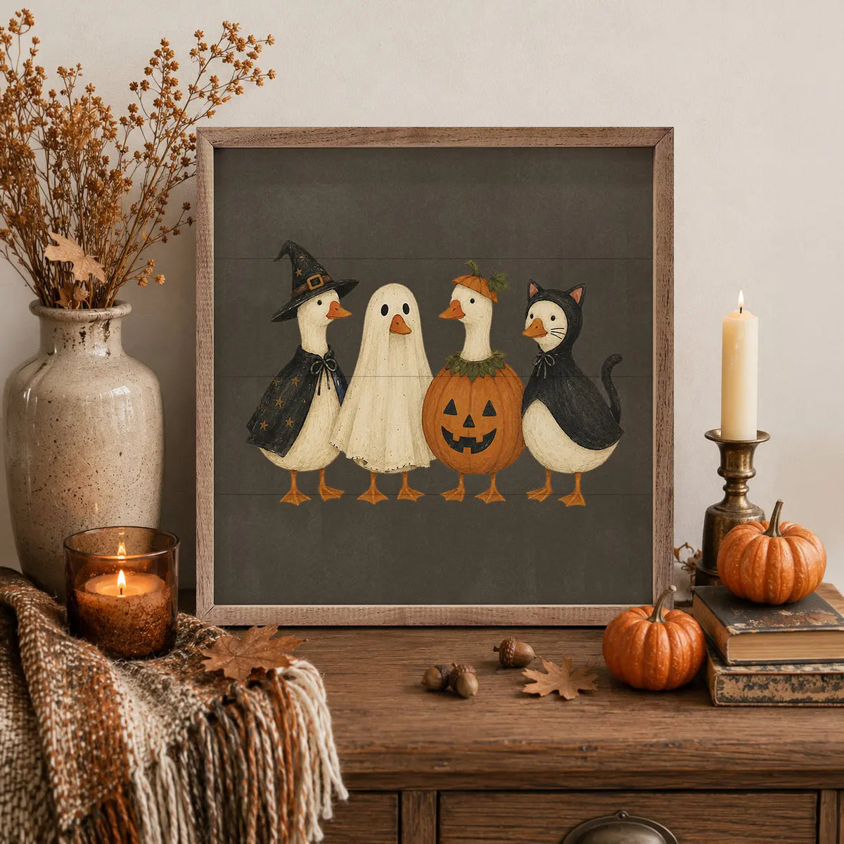 A framed Halloween art print featuring four whimsical goose characters in costumes (witch, ghost, pumpkin, and cat) on a dark gray background, styled on a wooden console table with a candle, pumpkins, acorns, and a vase of dried branches.