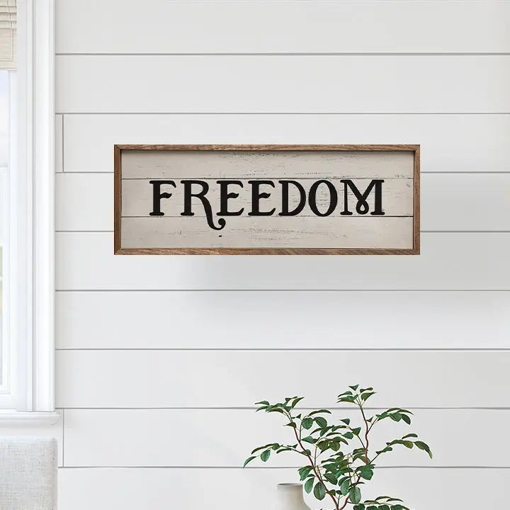 Wooden sign with 'FREEDOM' on a white wall with a plant in the corner