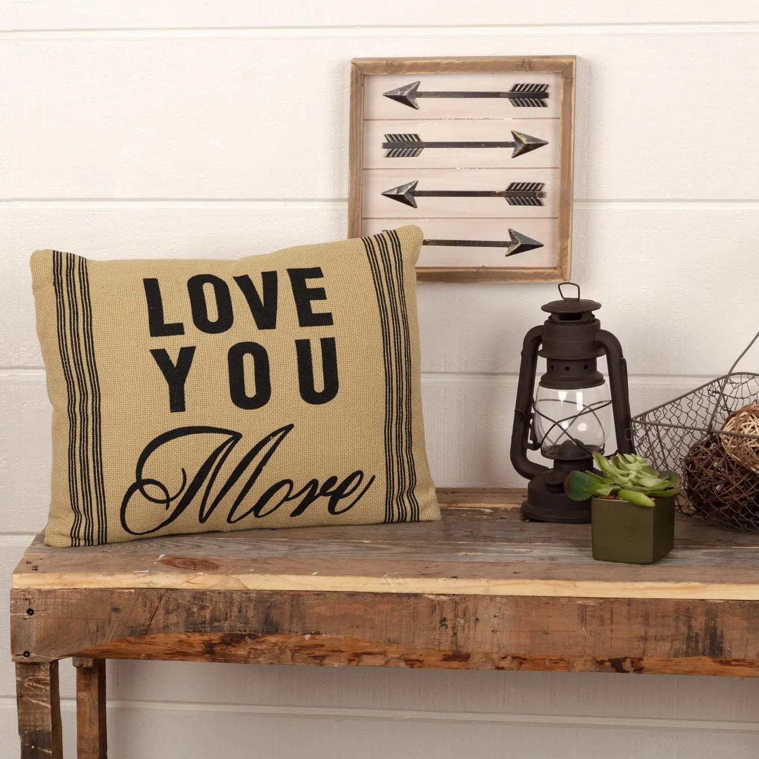 Cozy farmhouse pillow with 'Love You More' text, rustic wood bench, arrow wall art, lantern, and succulent decor