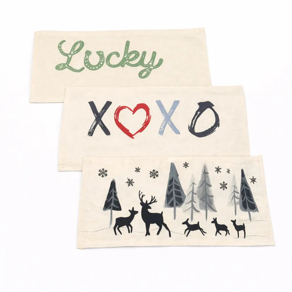 Three rectangular cream fabric napkins or placemats stacked diagonally on a white background. The top napkin has the word 'Lucky' in green script.