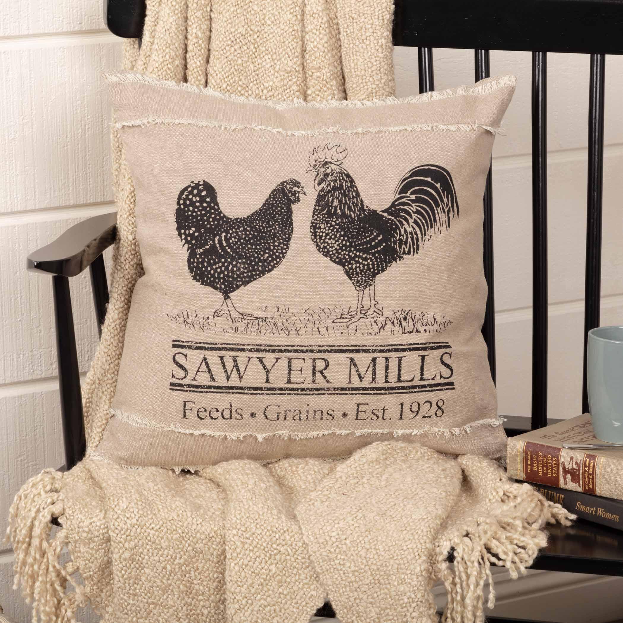 Farmhouse pillow with rooster and hen design, 'Sawyer Mills' text, frayed edges, on black chair with cozy blanket