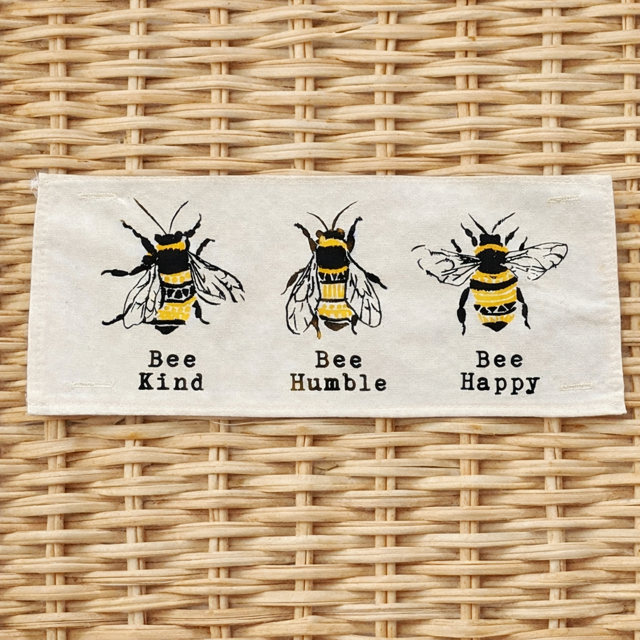 A rectangular cream-colored fabric patch featuring three black and yellow embroidered bees with the words 'Bee Kind', 'Bee Humble', and 'Bee Happy' printed below each. The patch is displayed flat on a light brown woven wicker background.