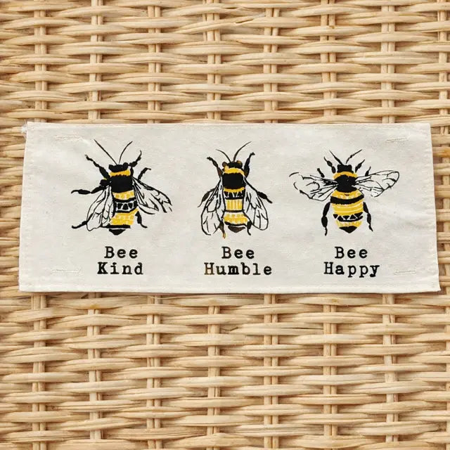 A rectangular cream-colored fabric patch featuring three black and yellow embroidered bees with the words 'Bee Kind', 'Bee Humble', and 'Bee Happy' printed below each. The patch is displayed flat on a light brown woven wicker background.