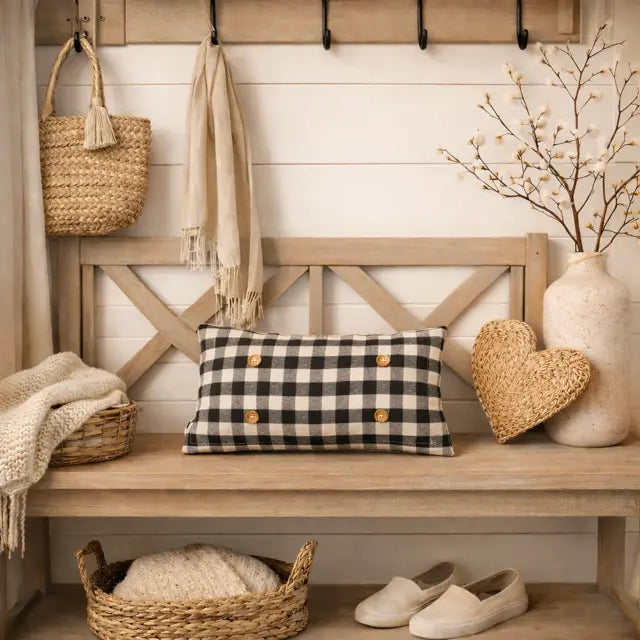 A rectangular black and beige gingham throw pillow with four gold buttons is styled on a rustic wooden bench in a cozy entryway setting, surrounded by home decor including a woven tote bag, a cream scarf, a woven heart-shaped decor piece, a vase with branches, and a pair of beige slip-on shoes.