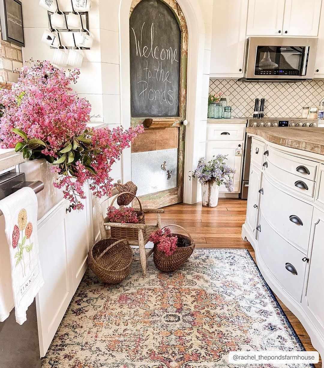 Cozy farmhouse kitchen with white cabinets, floral rug, wicker baskets, and pink blooms for cottage-style decor