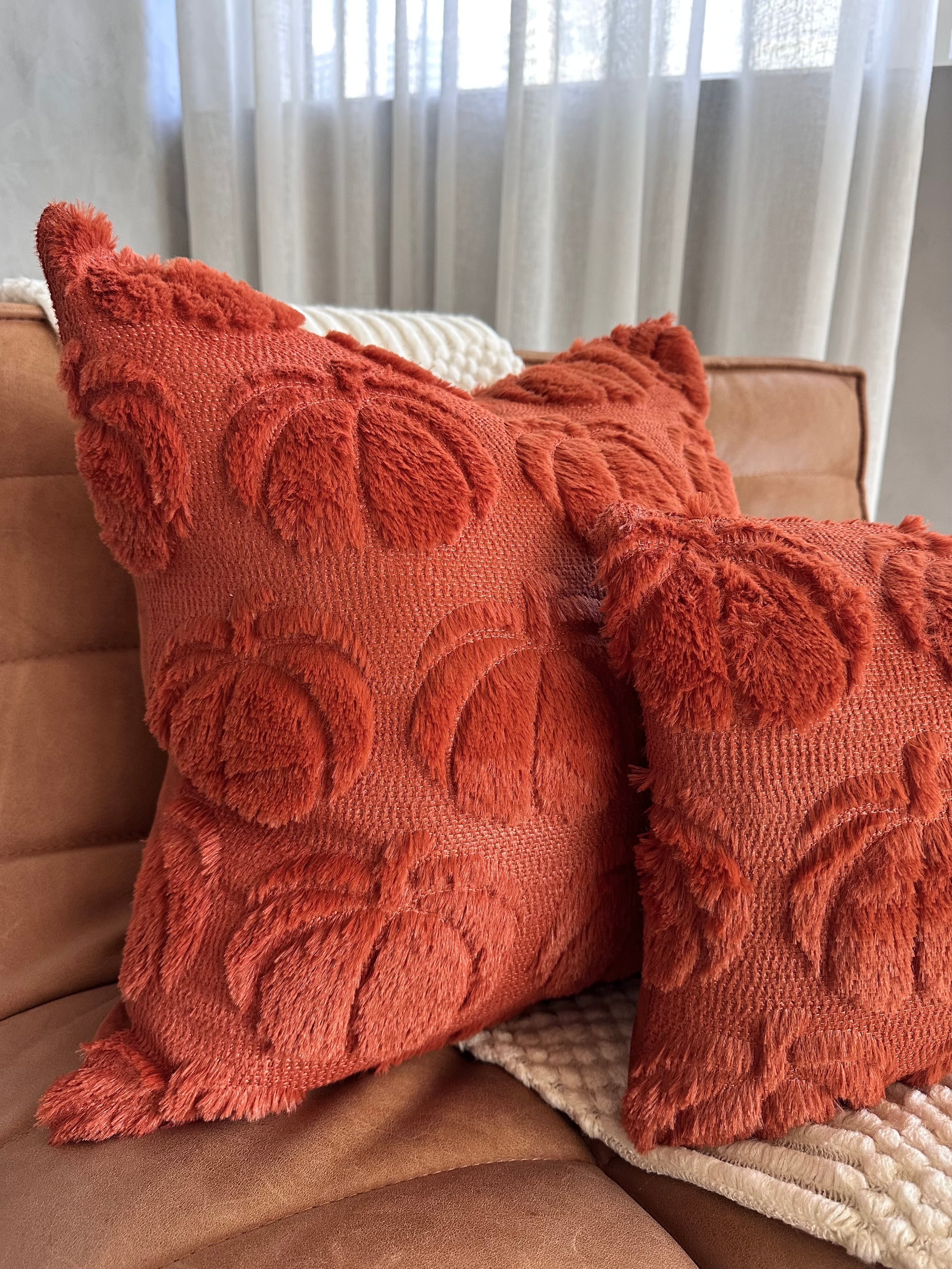 Two textured orange pillows on a brown couch with light curtains in the background.