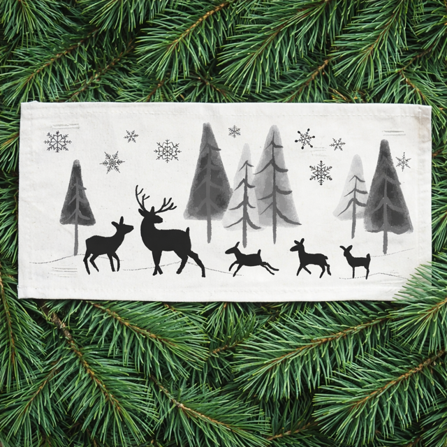 A rectangular white fabric napkin or tea towel featuring a black winter forest design with silhouettes of deer, pine trees, and snowflakes, laid flat on a bed of green pine branches.