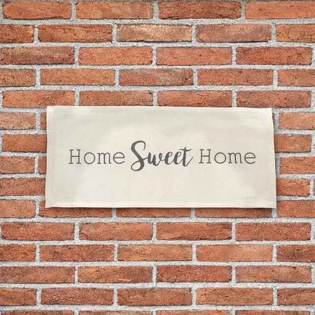 A rectangular beige canvas sign with the phrase 'Home Sweet Home' in dark gray lettering, mounted on a red brick wall.