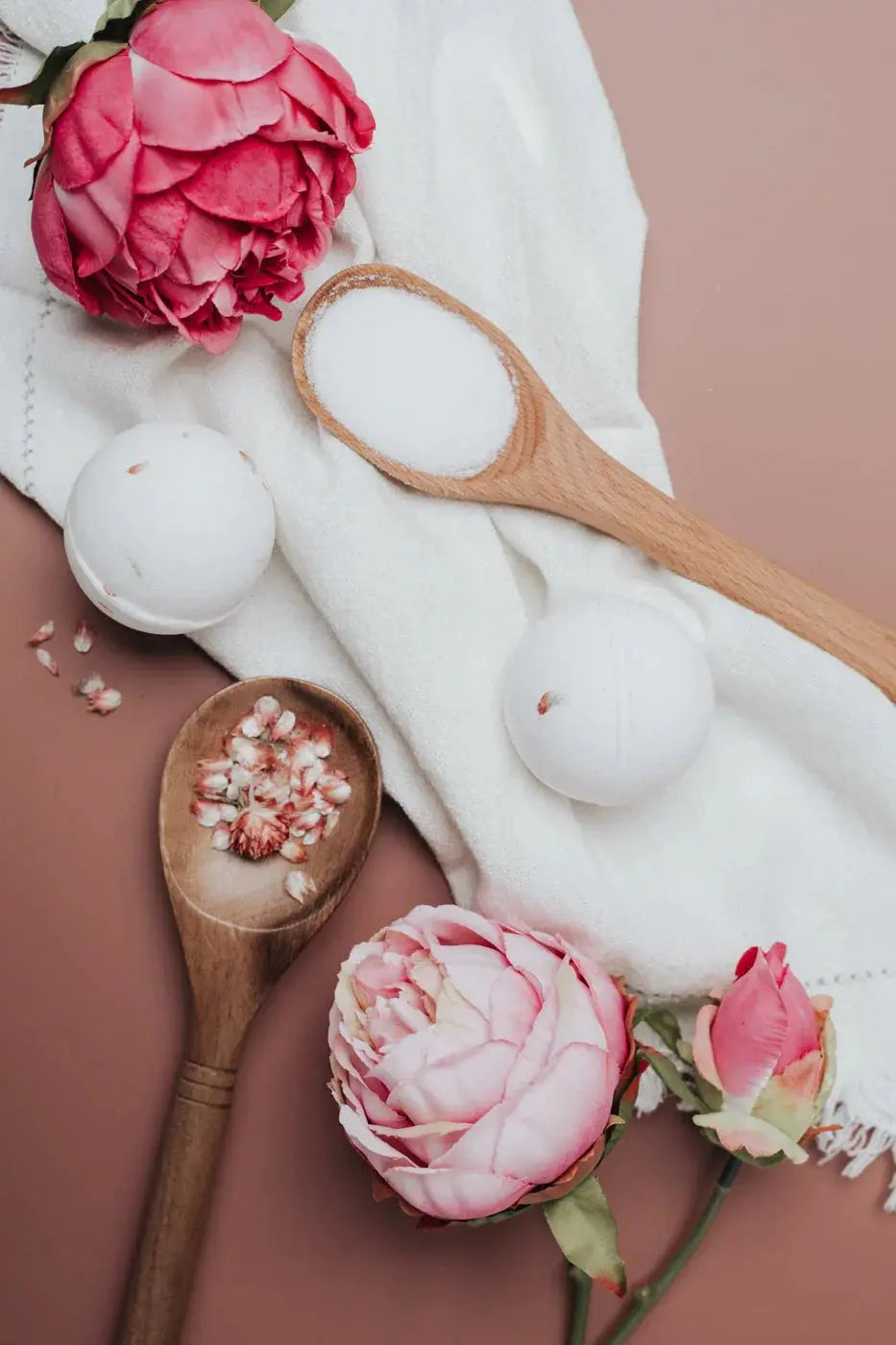 Cozy bath essentials with pink peonies, wooden spoons, and bath bombs on a soft towel