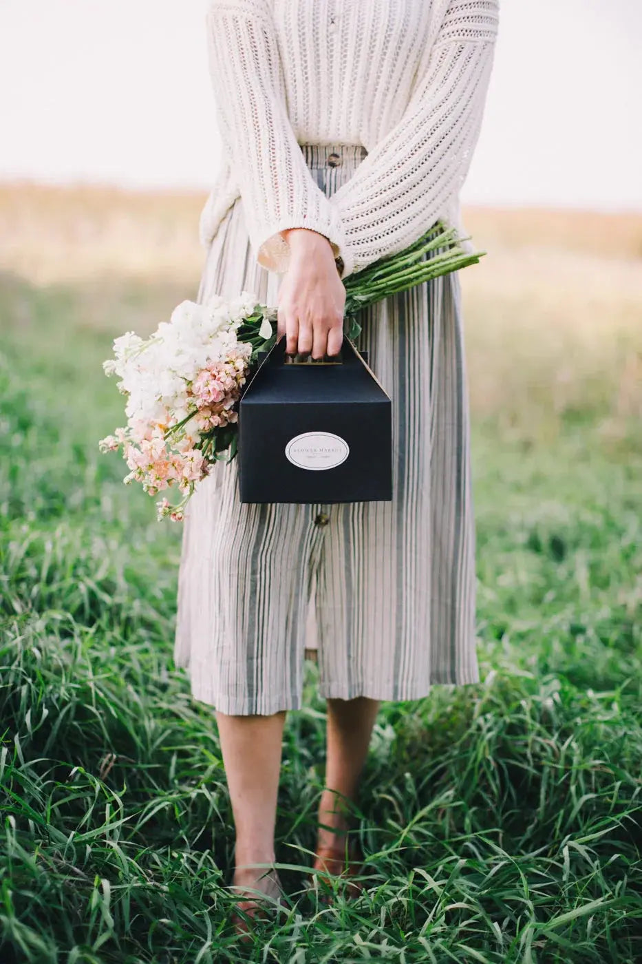 Woman holding fresh pink and white flowers in black box, standing in grassy field, cozy farmhouse style