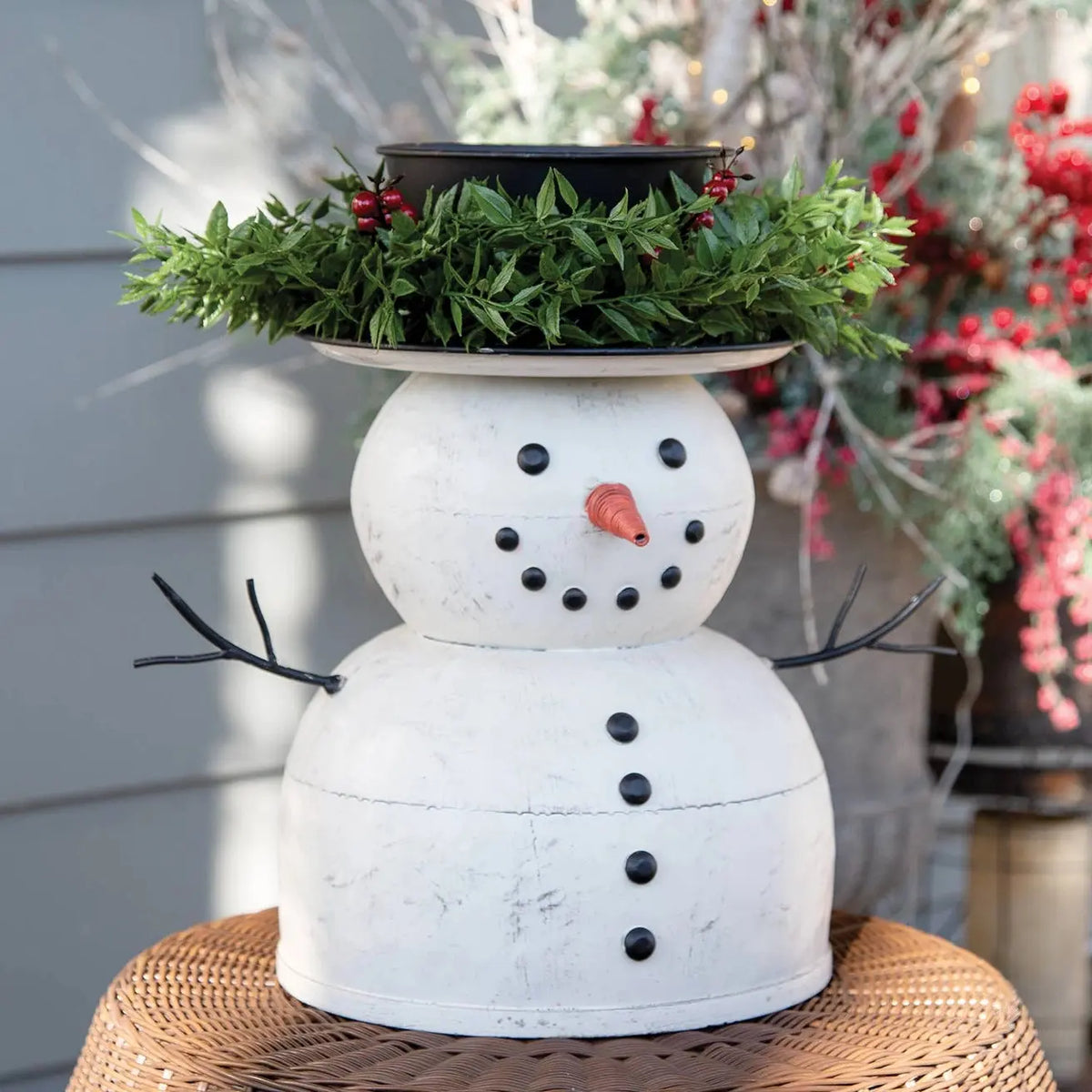 A white snowman planter with a distressed finish, featuring black buttons, a small orange carrot nose, and black metal arm branches. The planter is topped with a hat-shaped holder filled with green pine garland and red berries, displayed on a woven wicker stool against a gray siding background.