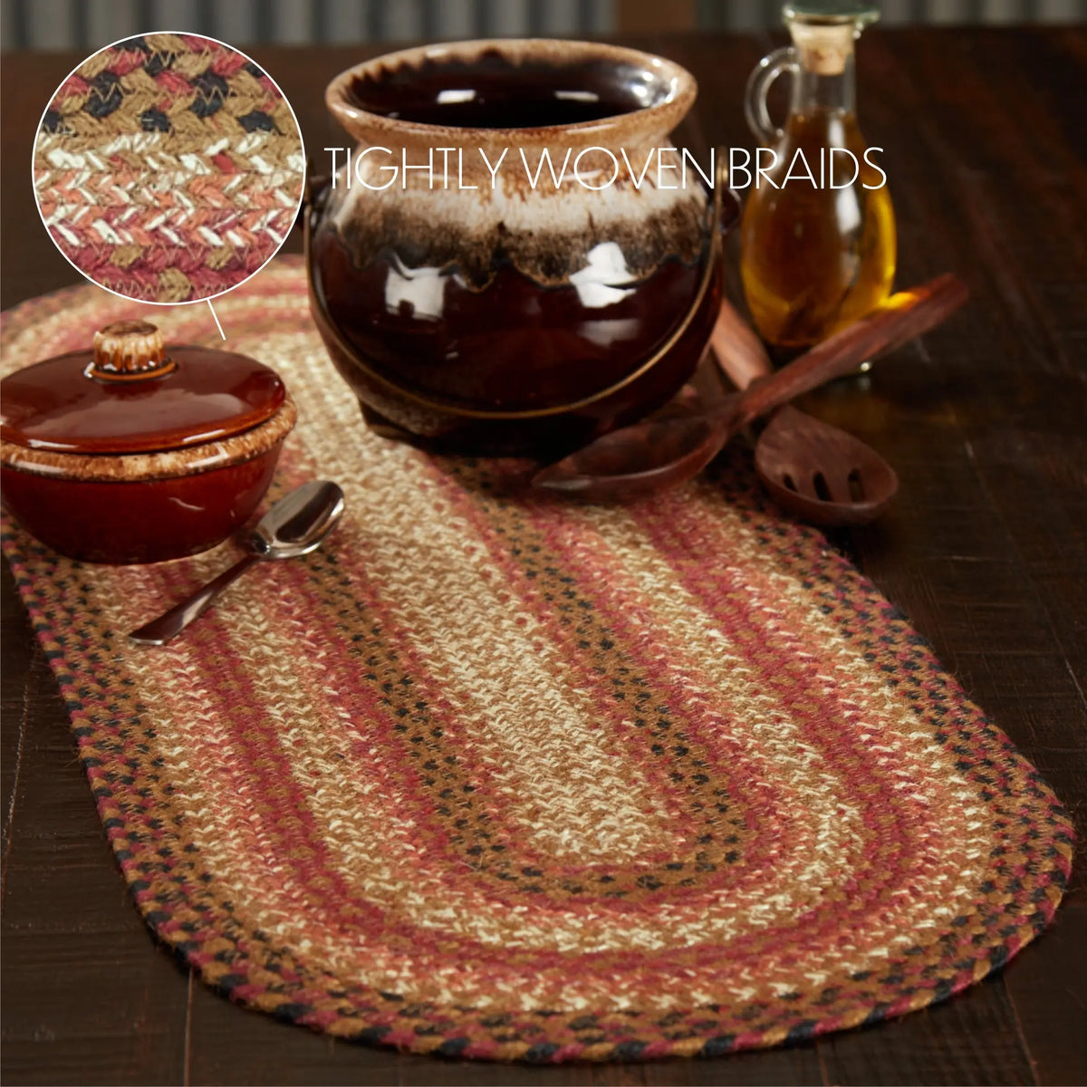 Braided oval table runner in a rustic multicolor pattern of beige, rust red, and brown, styled on a dark wooden table with ceramic dishes and kitchen utensils. A circular inset in the top left shows a close-up of the woven texture.