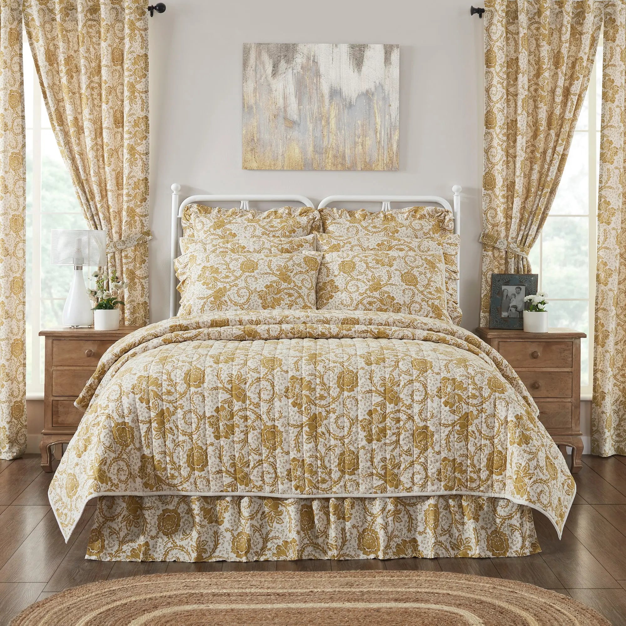 A bed dressed in a cream and mustard yellow damask-patterned comforter set with matching shams and a ruffled bed skirt, styled in a bright bedroom with matching curtains, wooden nightstands, and a wall art piece.