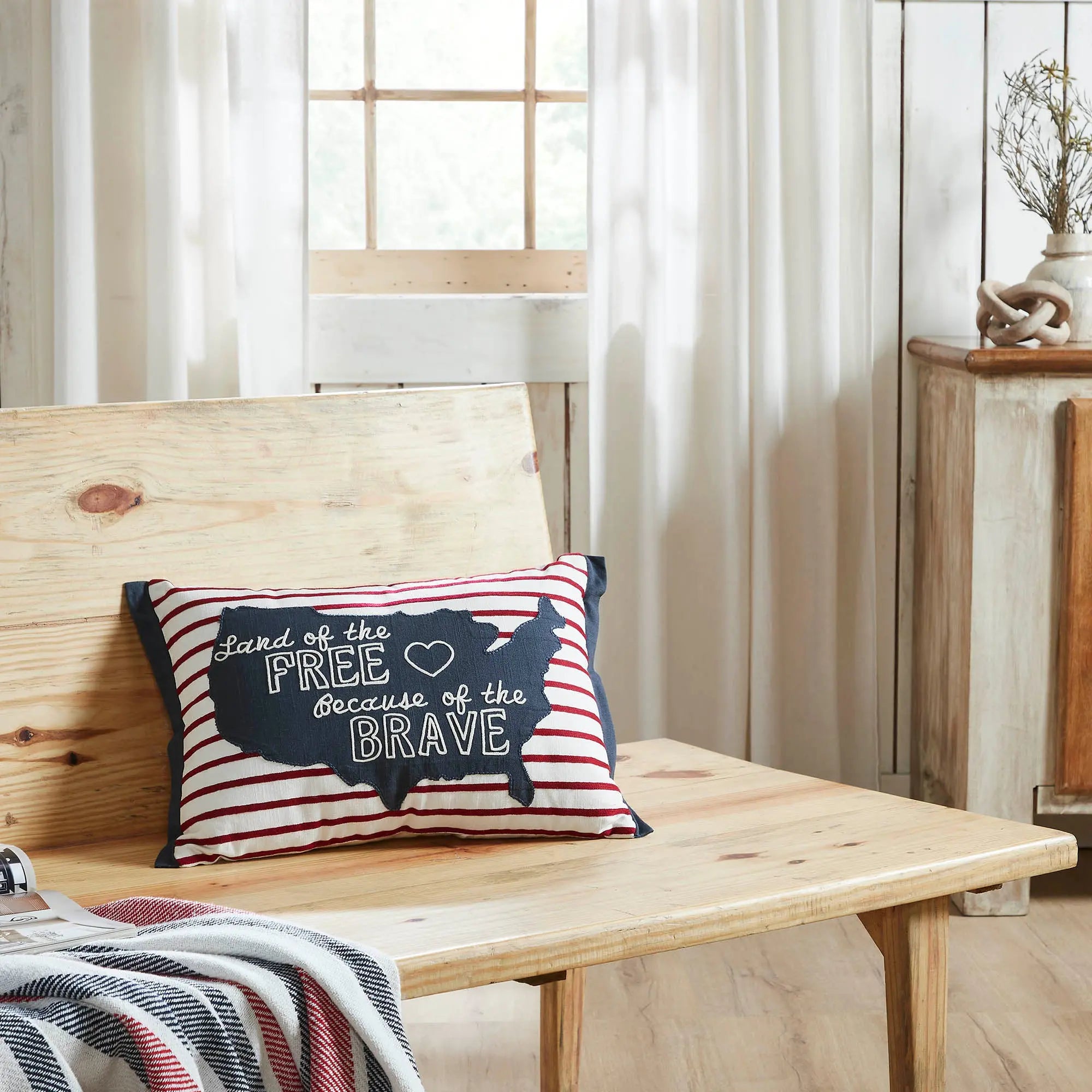 Decorative pillow with text on a wooden bench in a room with curtains and a side table.