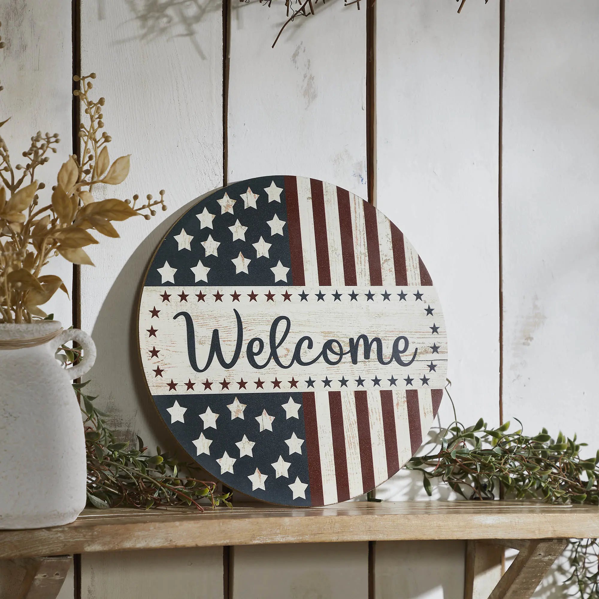 Decorative round sign with American flag pattern and 'Welcome' text on a wooden surface.