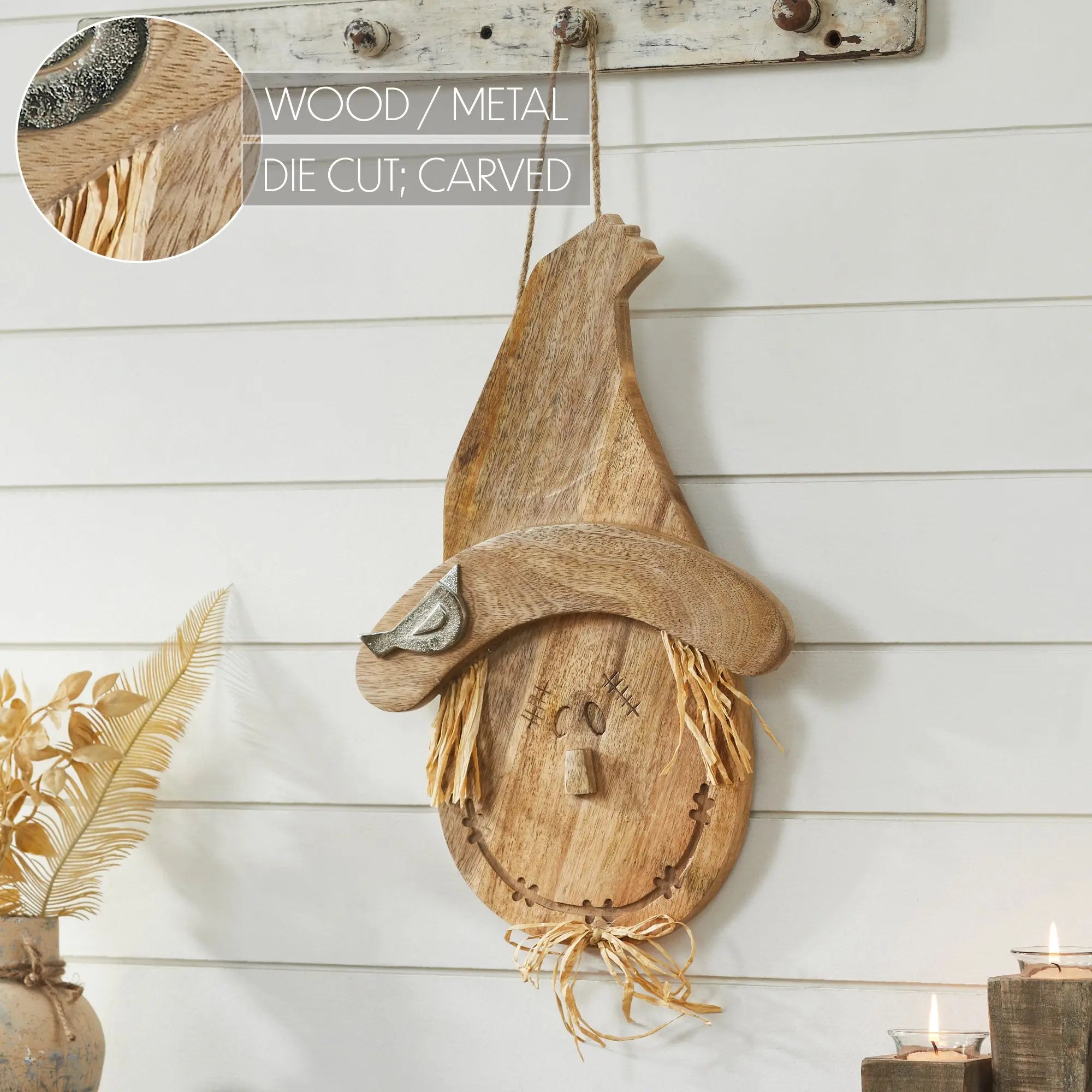 A rustic wooden scarecrow wall hanging with a carved face, straw hair, and a pointed hat, displayed against a white shiplap wall. A circular inset at the top left shows a close-up of the wood grain and die-cut detail, with text reading 'WOOD / METAL DIE CUT; CARVED'.