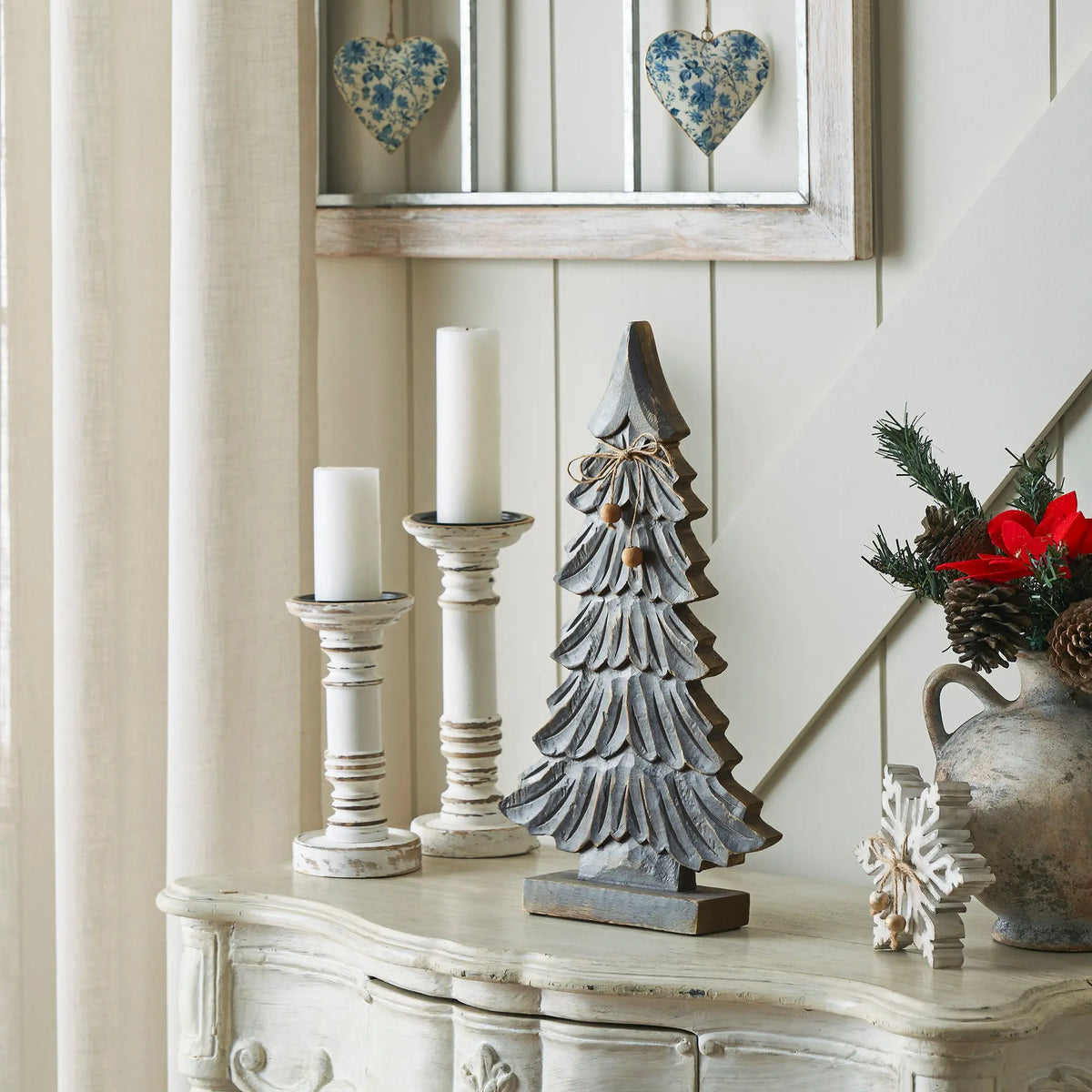 A rustic wooden Christmas tree ornament stands on a distressed white console table in a styled holiday interior. The ornament has layered scalloped edges and a small bow with beads at the top.
