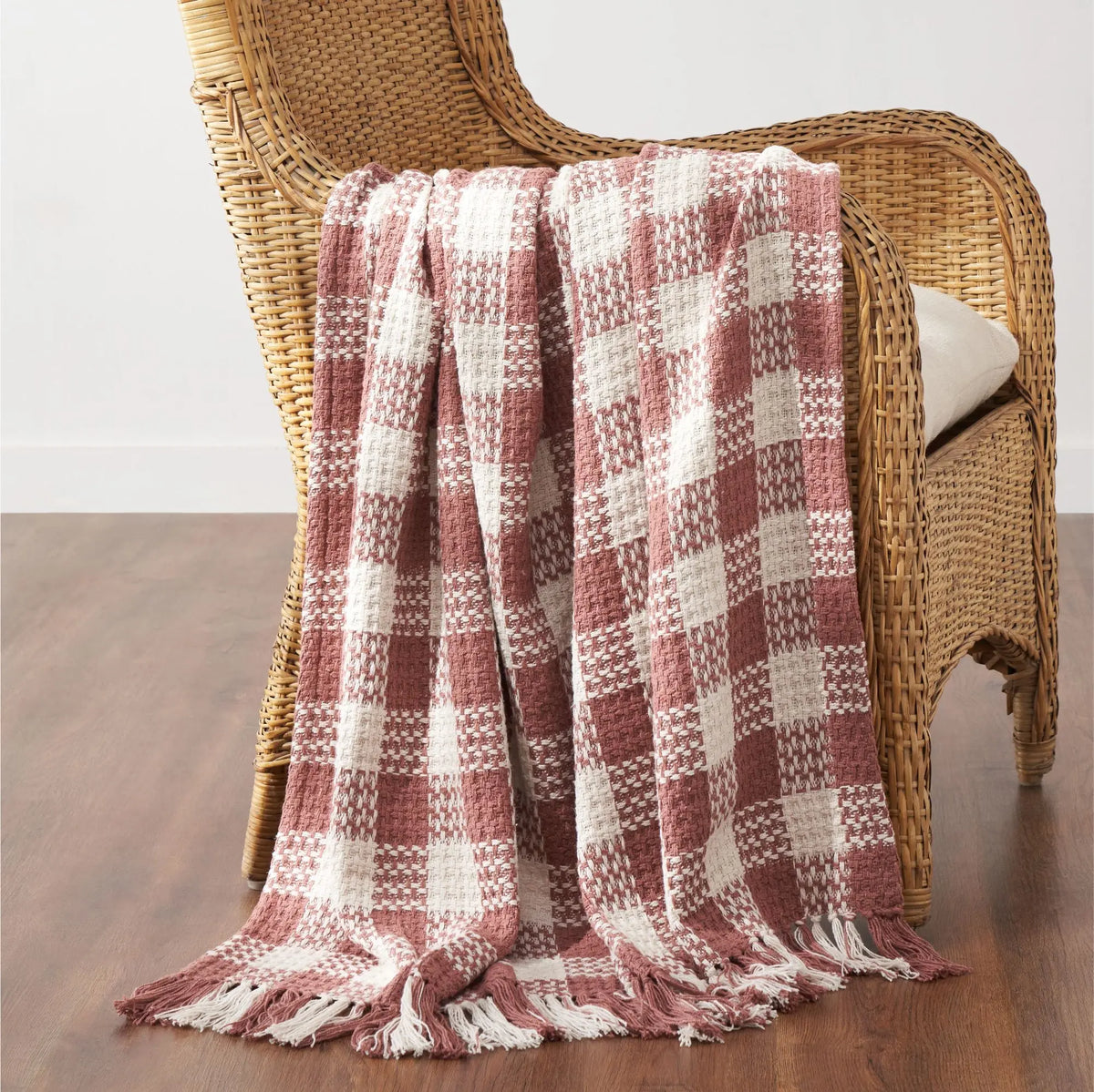 A woven throw blanket with a dusty rose and cream plaid pattern and fringe edges, draped over a wicker armchair on a wooden floor.