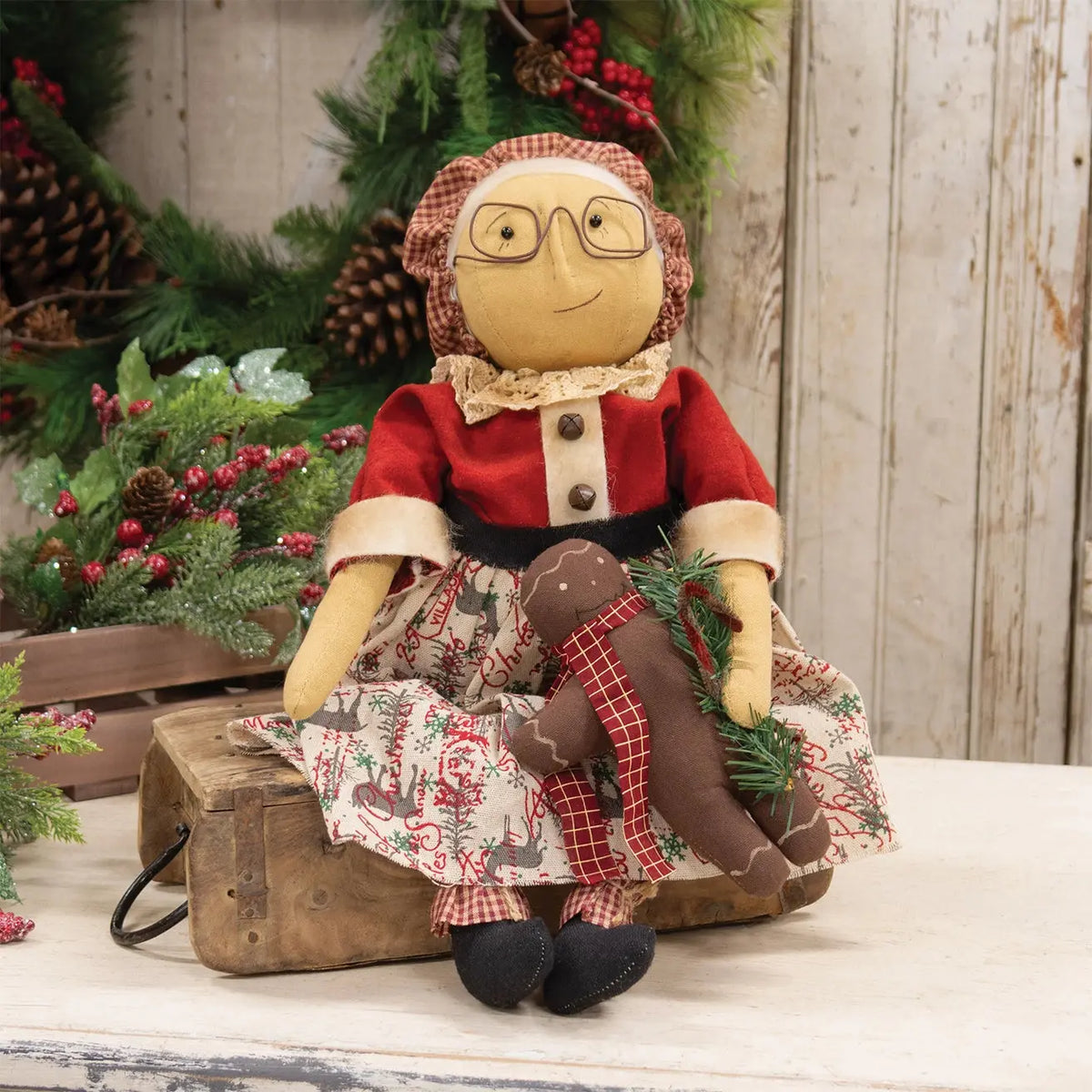 A rustic Christmas doll with a red dress, patterned skirt, and brown plaid bonnet sits on a wooden trunk, holding a smaller brown doll, with a holiday wreath in the background.