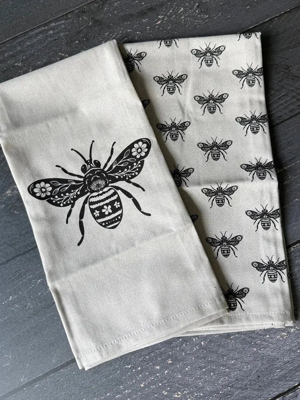 Two white towels with black bee patterns on a dark wooden surface