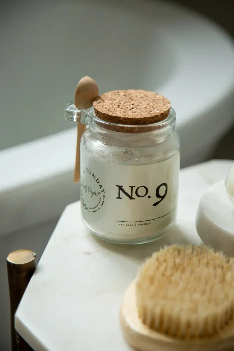 Glass jar of No. 9 bath salts with cork lid, wooden spoon, and natural bristle brush on marble, farmhouse bathroom decor