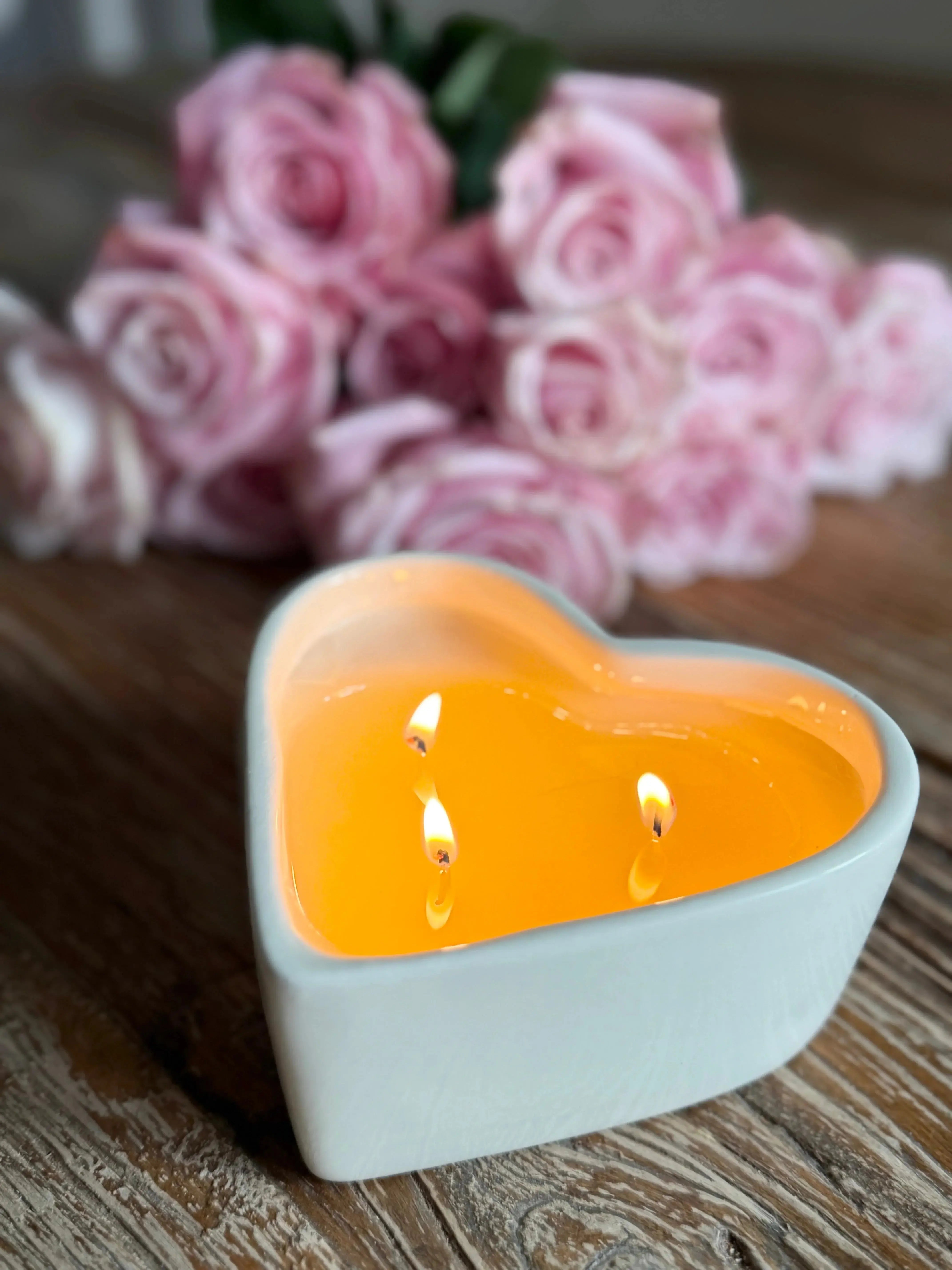 Heart-shaped candle with three wicks, glowing warmly beside pink roses on rustic wood