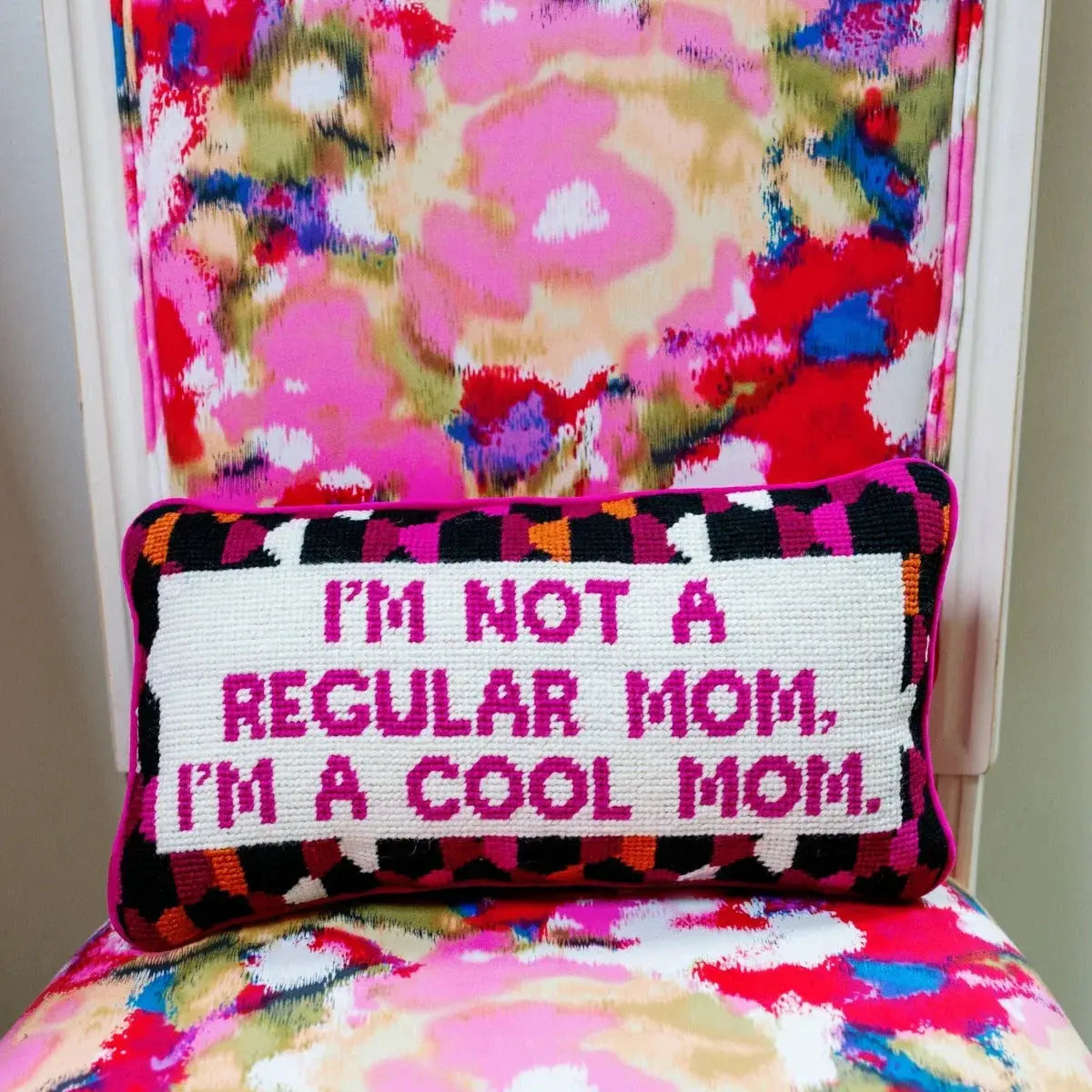 Fun needlepoint pillow saying 'I'm not a regular mom, I'm a cool mom' on colorful floral chair, cozy home decor