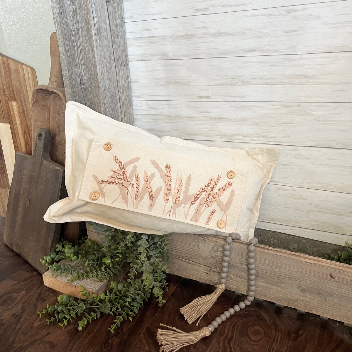 A rectangular cream throw pillow with embroidered wheat stalks and decorative buttons, styled on rustic wood planks with a beaded tassel and greenery.