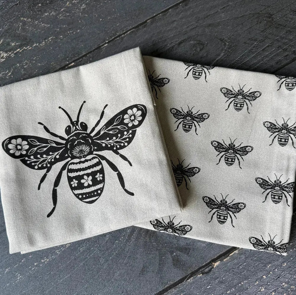 Two folded cream-colored linen napkins with black printed bee designs, displayed on a dark gray wooden surface.