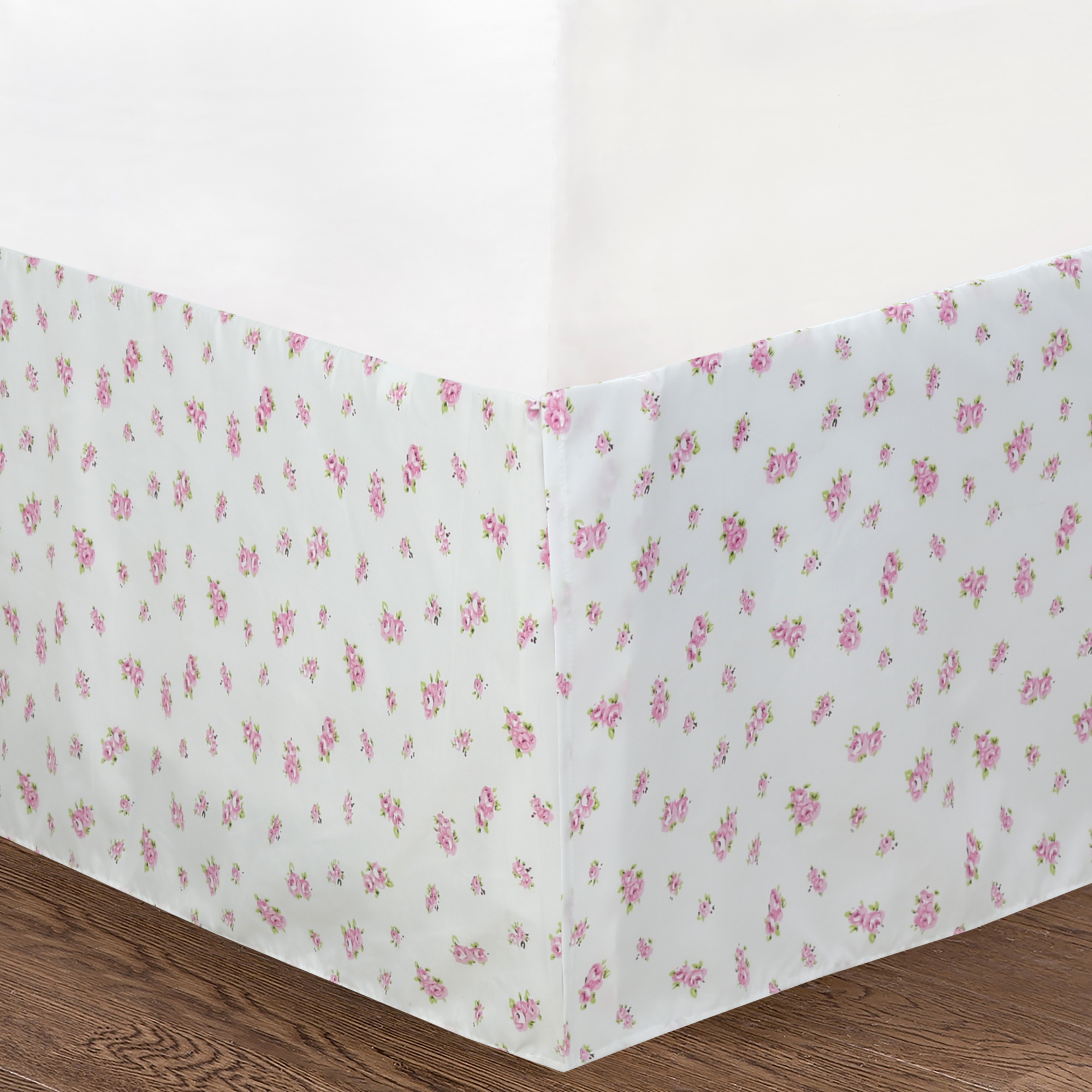 A white bed skirt with a small pink floral pattern, shown fitted around the corner of a bed on a wooden floor.
