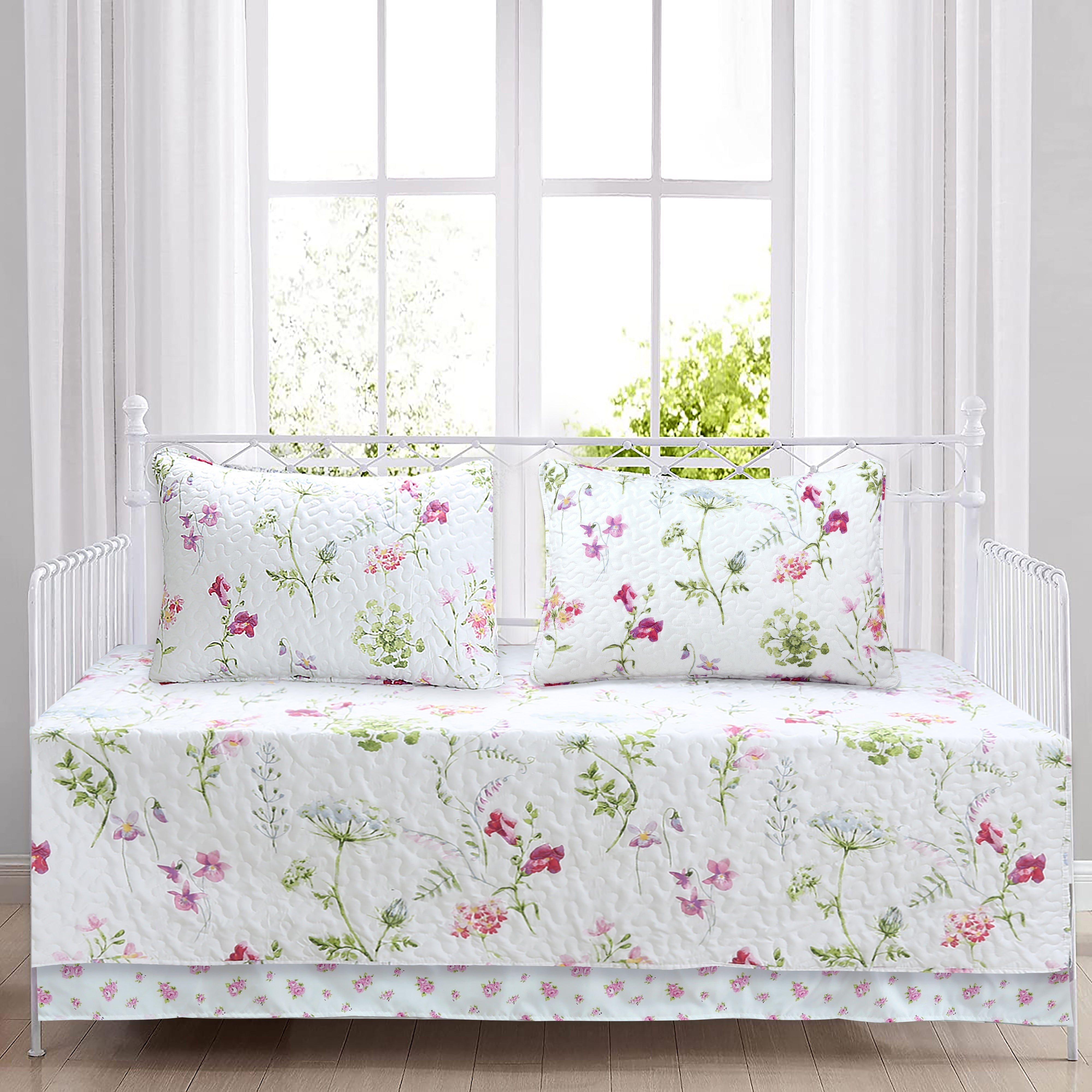 A white floral bedding set including a quilted coverlet, matching pillow shams, and a ruffled bed skirt, styled on a white metal daybed in front of a bright window.