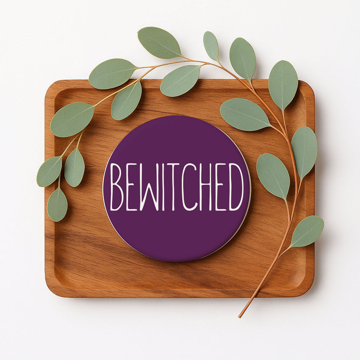 Wooden tray with a purple 'BEWITCHED' coaster and green leaves on a white background