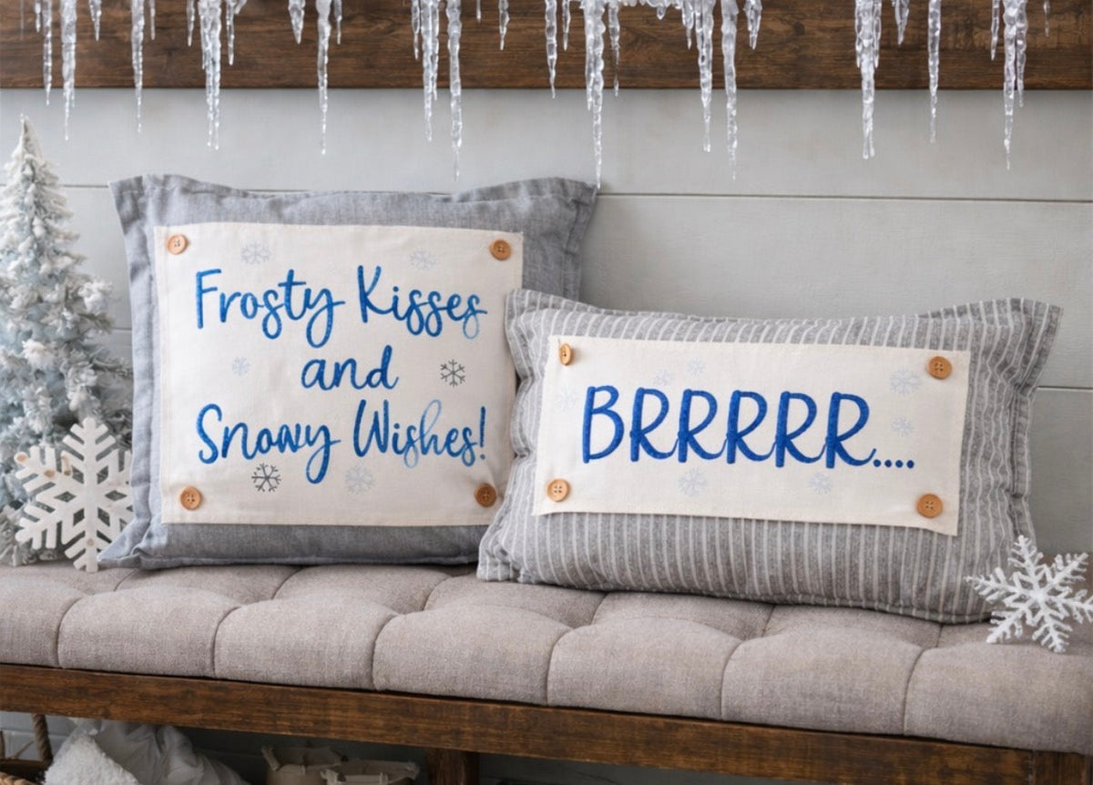 Two decorative throw pillows displayed on a tufted bench in a styled holiday setting. One pillow is light gray with a cream patch reading 'Frosty Kisses and Snowy Wishes!' in blue script, and the other is gray with a cream patch reading 'BRRRRR.