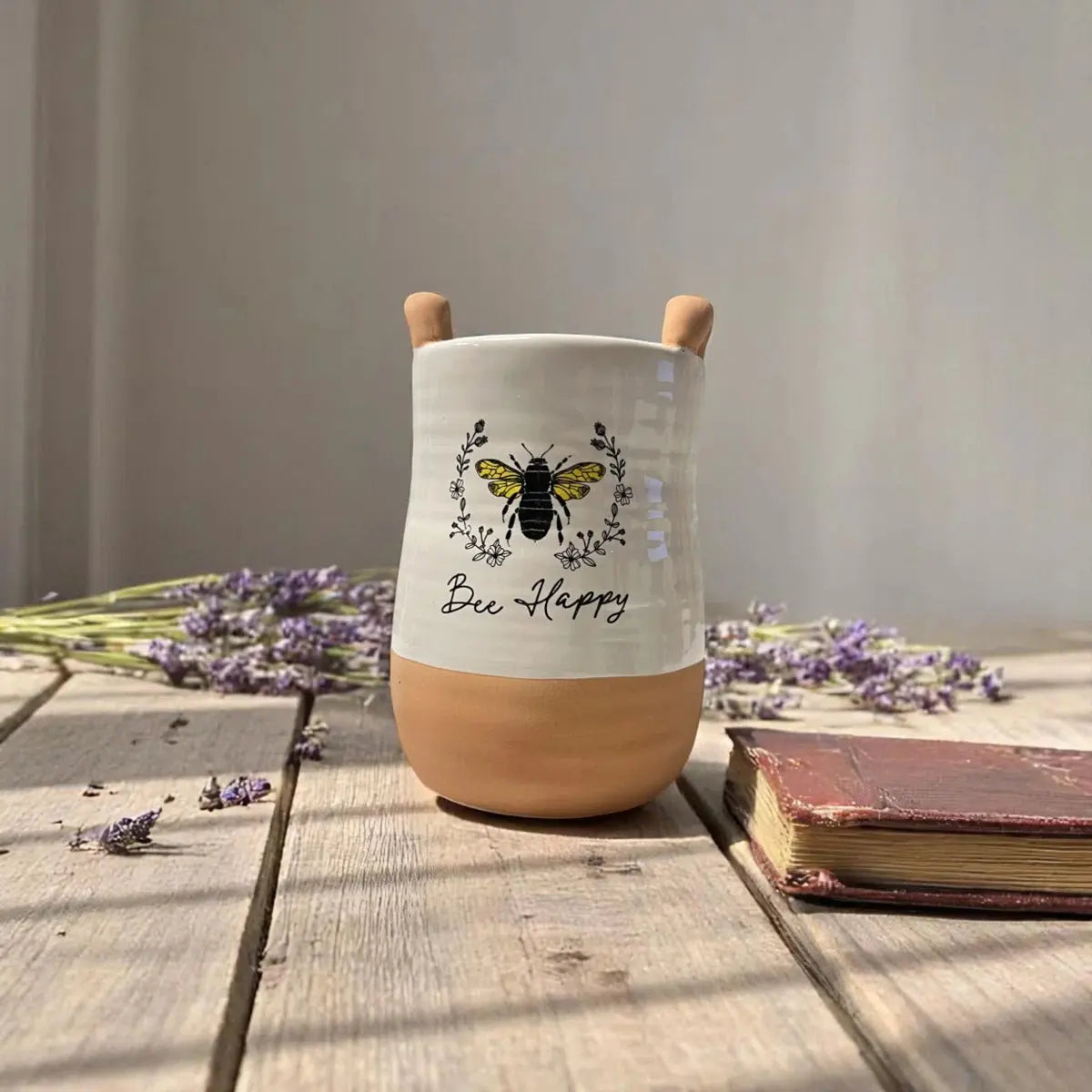 Ceramic mug with wooden handle and 'Bee Happy' text on a wooden surface with lavender and a book.