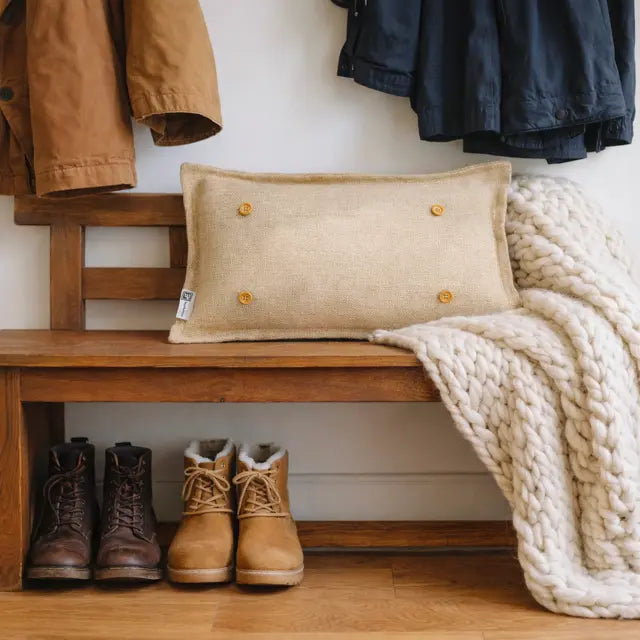 A rectangular beige linen throw pillow with four gold buttons, styled on a wooden bench next to a chunky cream knit blanket, with boots and jackets in the background.