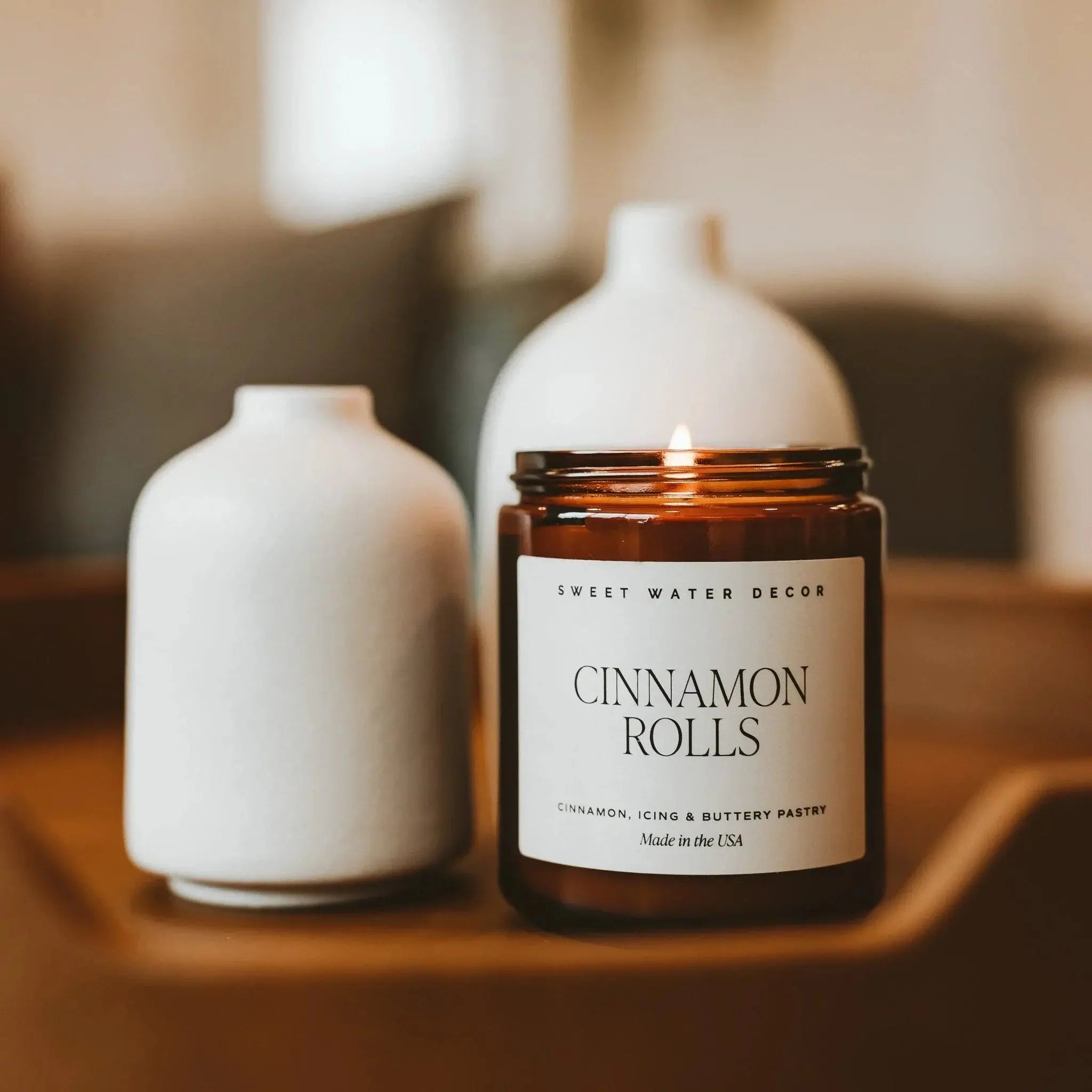 Candle labeled 'Cinnamon Rolls' by Sweet Water Decor with two white vases on a wooden surface.
