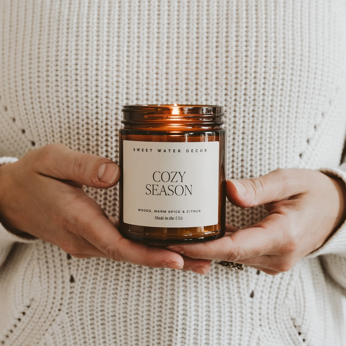 Person holding a 'Cozy Season' candle in front of a textured beige background