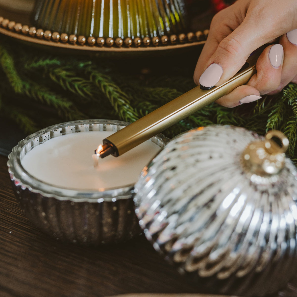 A close-up lifestyle shot of a hand with pale pink manicured nails lighting a white candle in a ribbed glass holder using a sleek gold metal lighter.